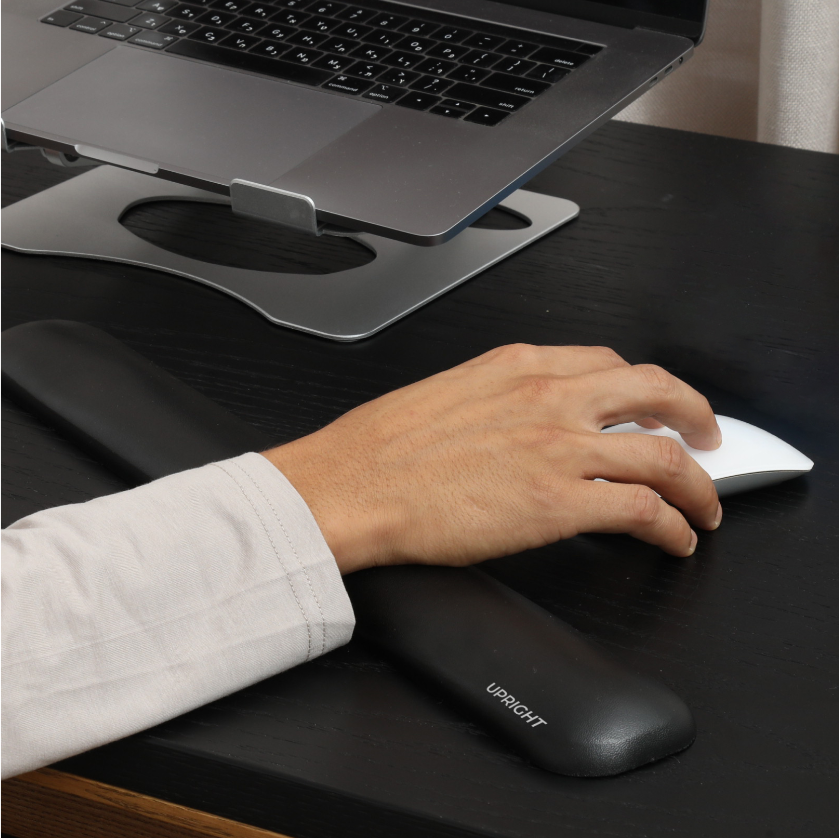 A person's hand using a black wrist rest while typing on a keyboard, with a laptop and mouse on a desk.